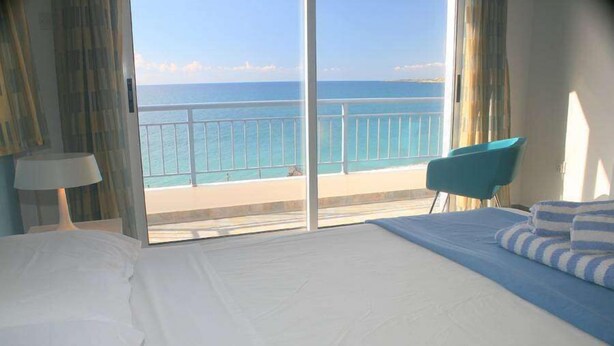 A hotel room with a large bed, a view of the sea, a balcony with a railing, and a blue chair. - Blue Coral Beach Villas (Paphos)