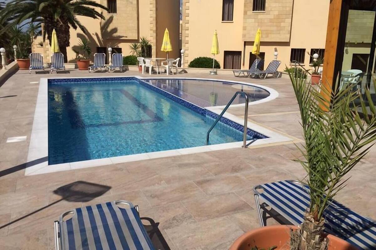 Une piscine entourée de chaises longues et de parasols, elle-même bordée de bâtiments à la façade beige.