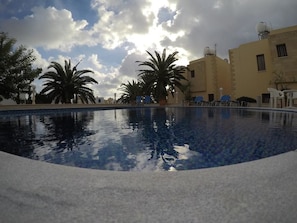 A swimming pool with clear water, surrounded by palm trees and a building with satellite dishes. - Dimma Seaside Houses (Chlorakas)