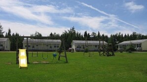 A grassy outdoor area with a playground, including swings and a slide, and a building with a covered porch in the background. - Joyce's Motel & Cottages (St. Peters)