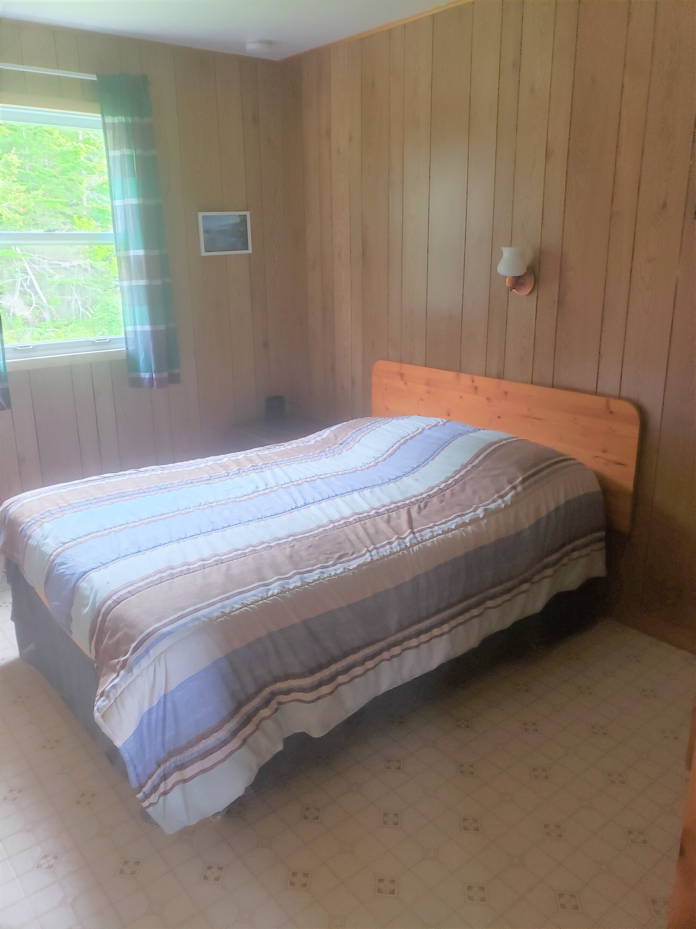 Un dormitorio con cabecera de madera, cama con ropa de cama a rayas, ventana con cortinas y una lámpara fijada a la pared.