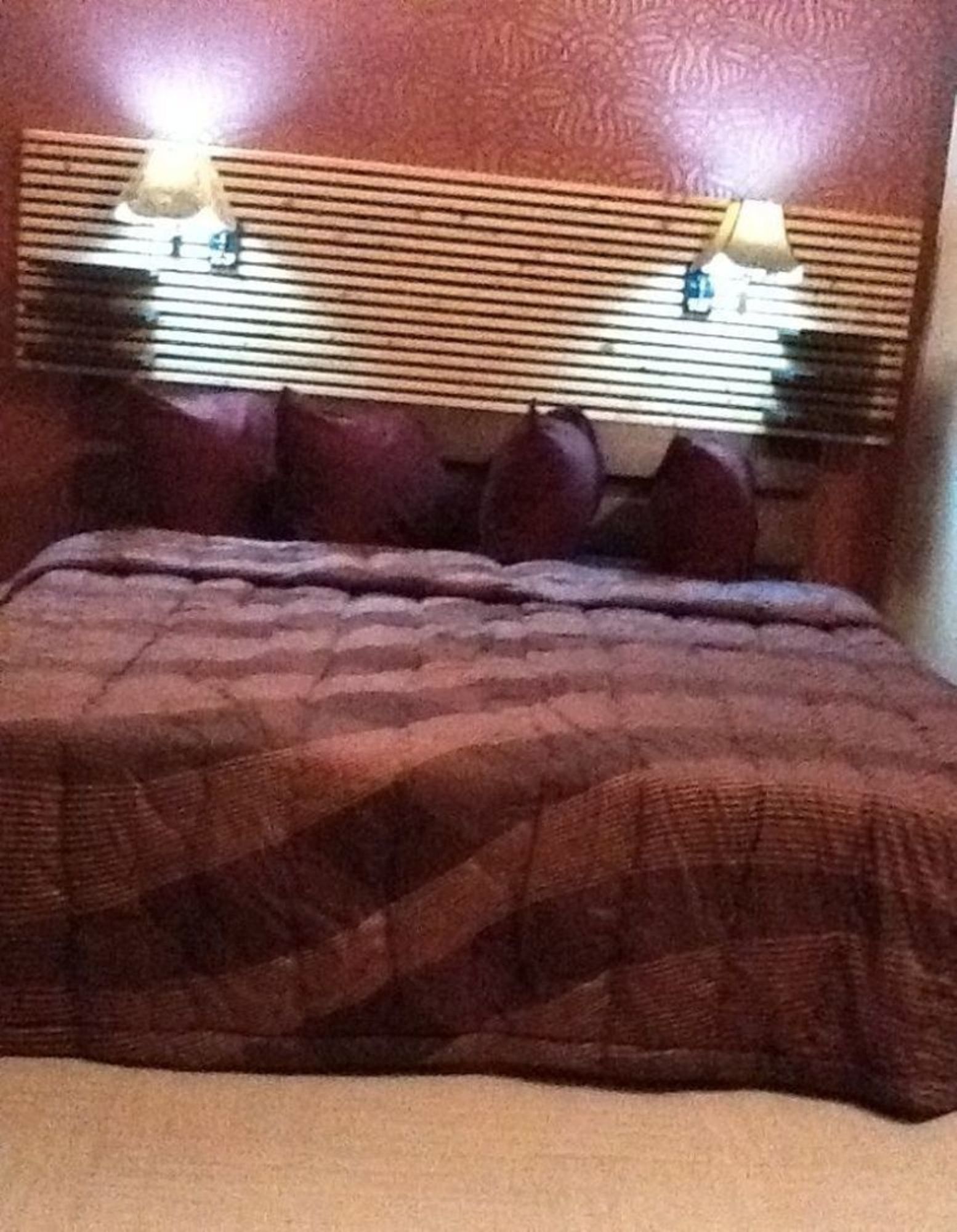 Una cama con una colcha de diseño oscuro, un cabecero con listones horizontales y dos lámparas fijadas a la pared.