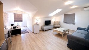 Ein modernes Wohnzimmer mit Sofa, Fernseher, kleinem Tisch und Sessel. Es gibt eine Küchenzeile mit Kühlschrank und Mikrowelle. Der Raum hat eine schräge Decke und eingelassene Beleuchtung.