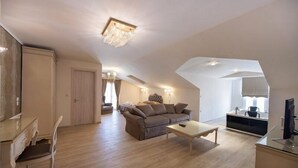 Ein großzügiger Wohnbereich mit Sofa, Couchtisch und Fernseher. Eine Tür führt in einen anderen Raum, ein Fenster mit Vorhängen.