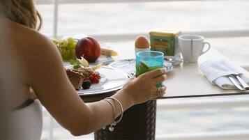 Una persona sentada a una mesa con alimentos para el desayuno, como fruta, huevos y una bebida verde, con vistas a un paisaje pintoresco.