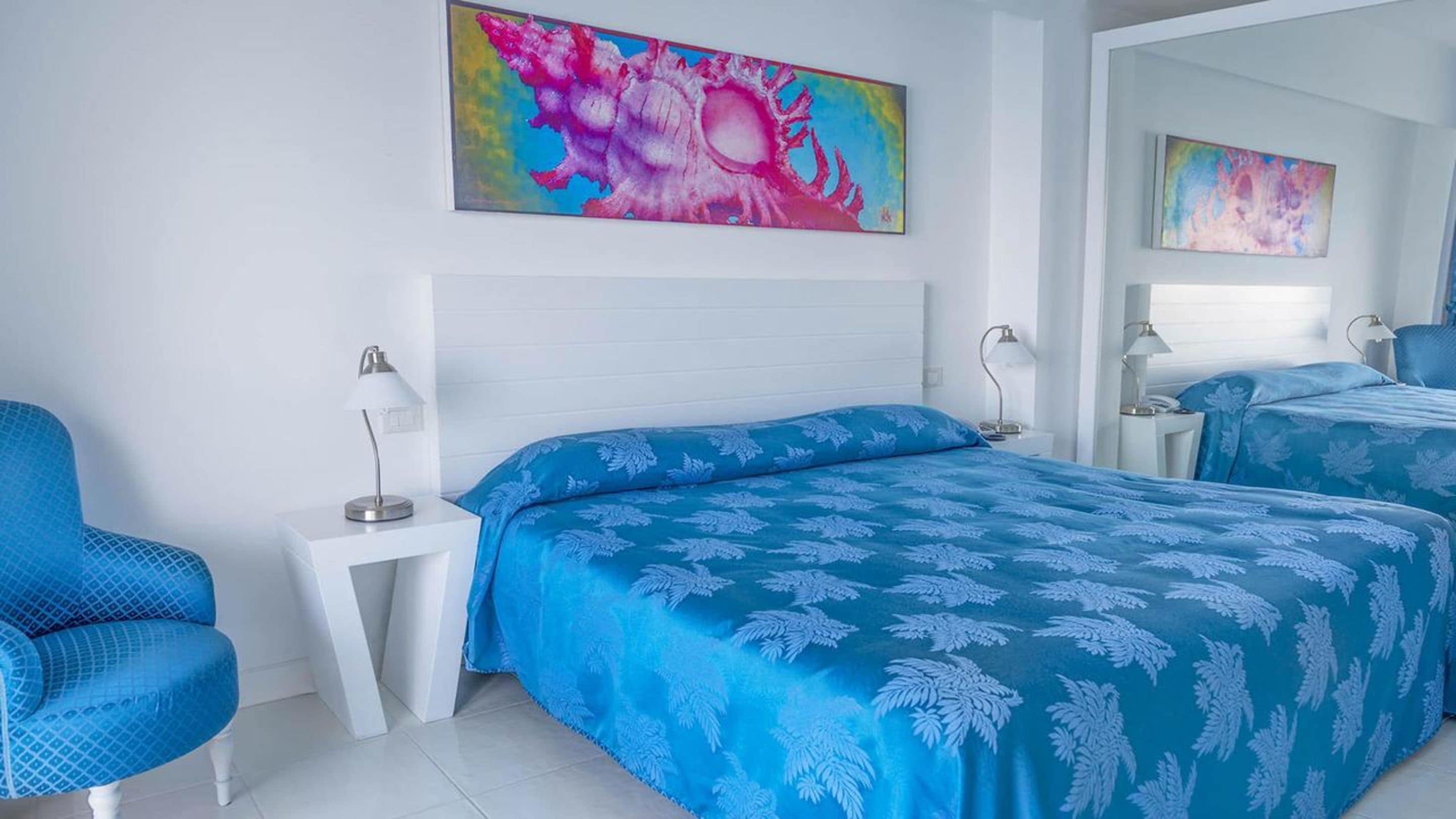 A hotel room with a large bed, a chair, a nightstand, and two paintings on the wall.