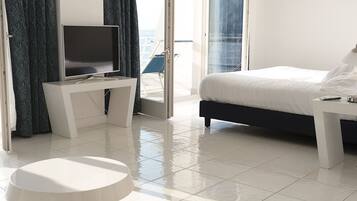 A modern hotel room with a bed, a TV on a white console table, a small round table, and a view of the sea through a sliding glass door.