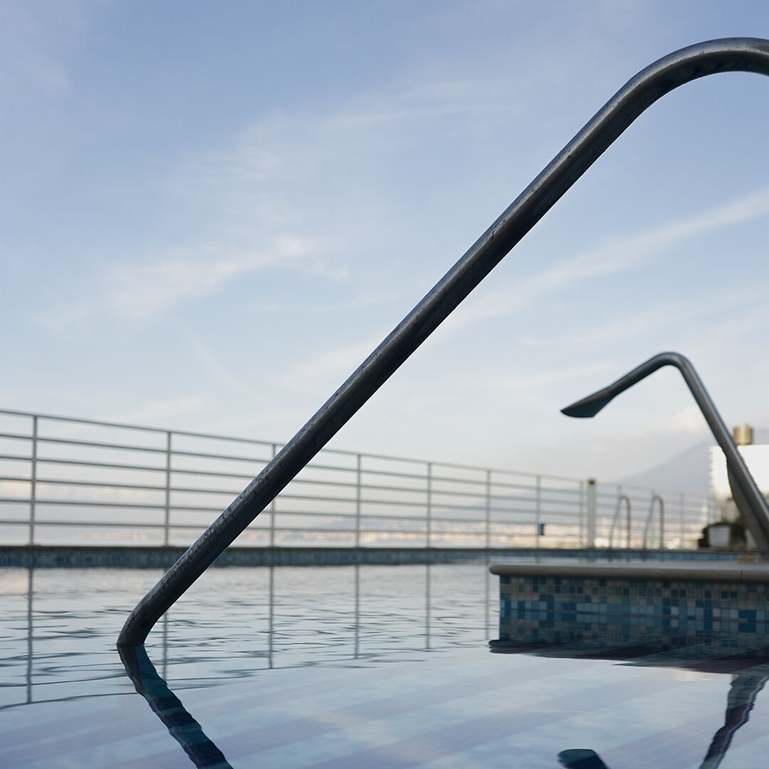 A metal pool ladder extending over a pool with a tiled edge and a metal railing in the background.