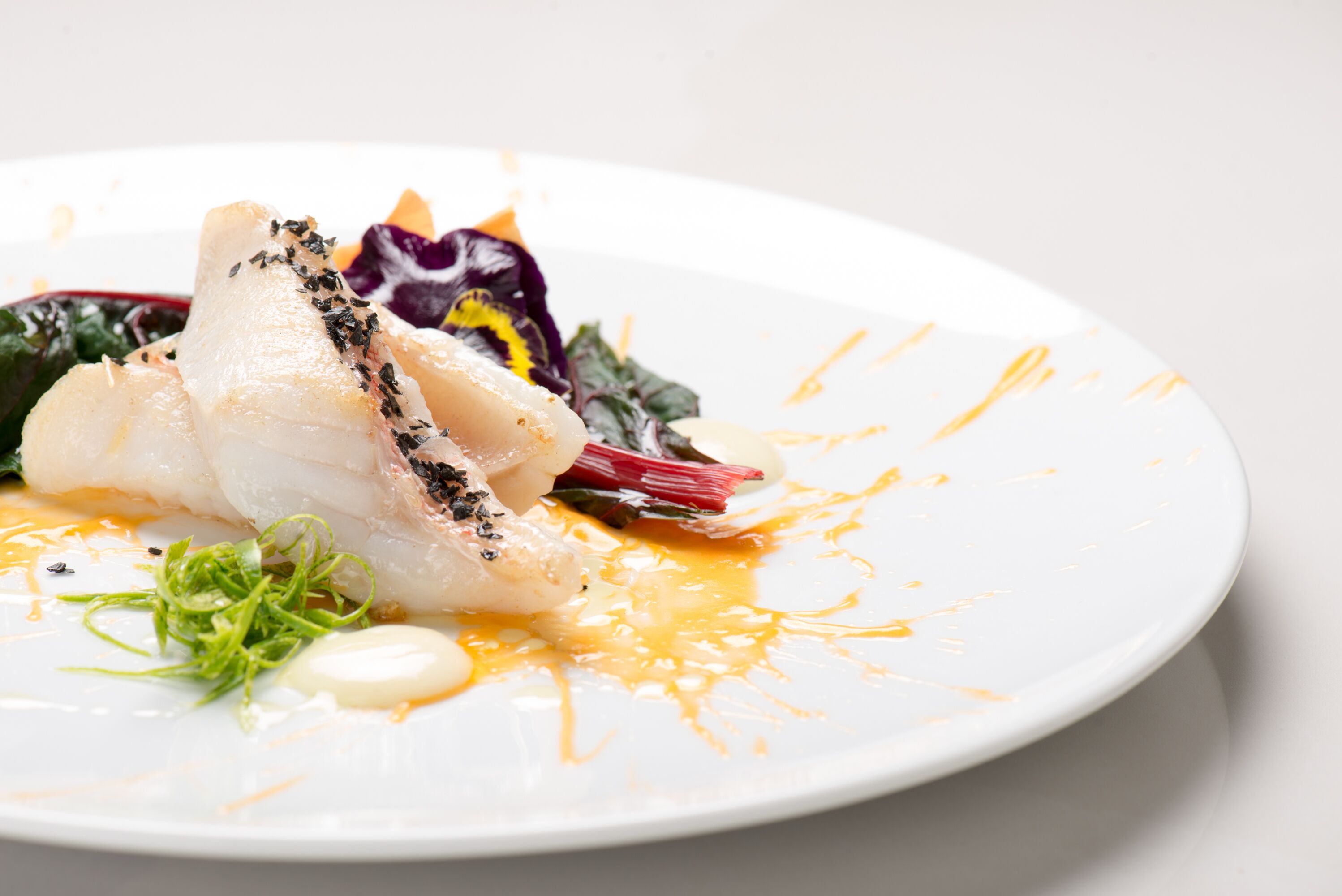 Un plato blanco con un trozo de pescado blanco, vegetales verdes y salsa amarilla.