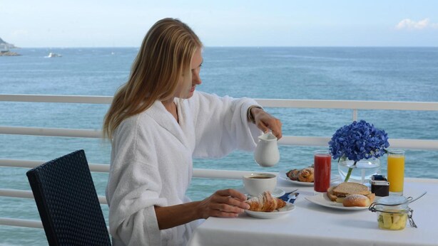 Una persona serviendo café en una taza sobre una mesa con alimentos para el desayuno, un jarrón de flores y vista al mar.