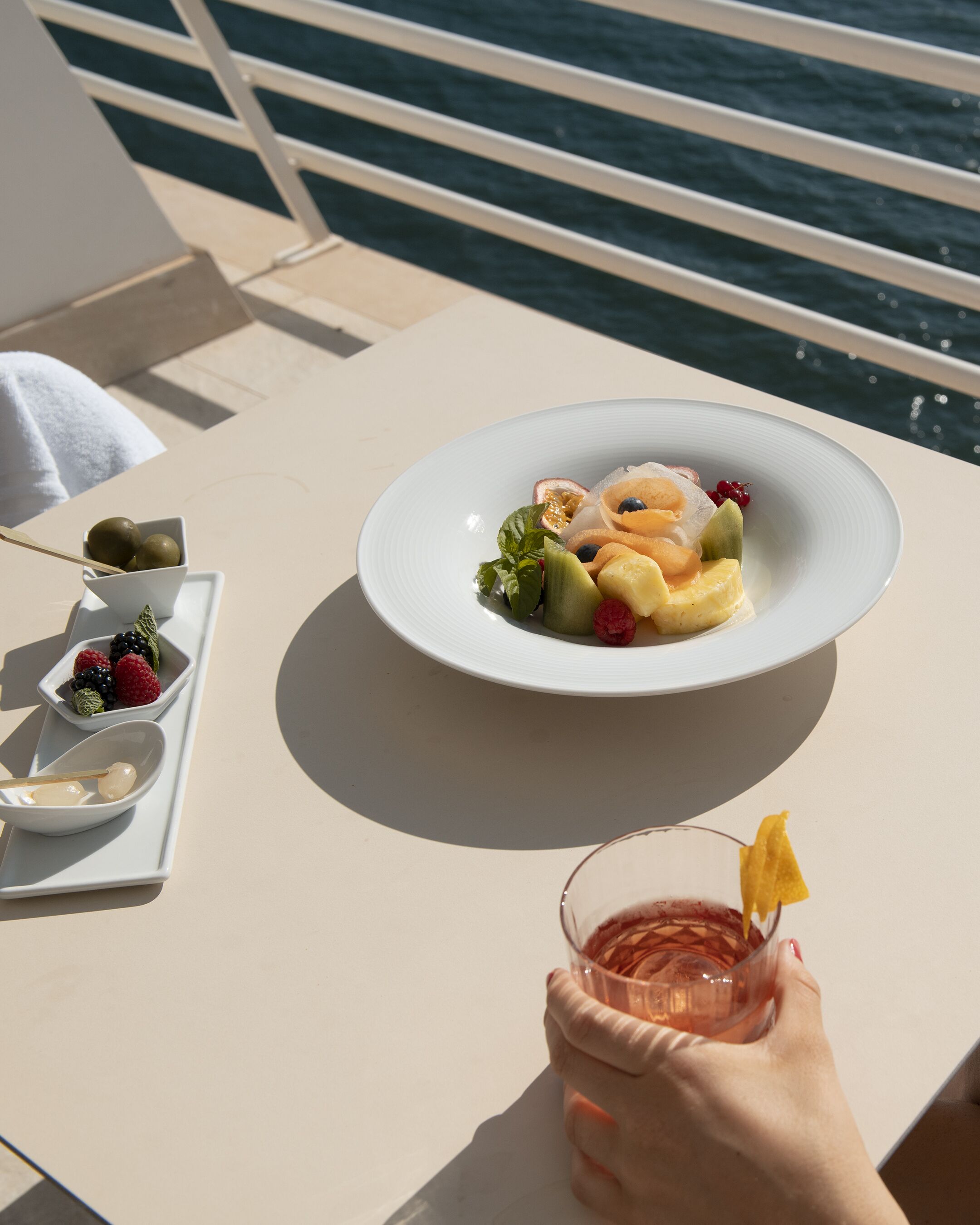 A table with a plate of fruit and a glass of drink, a bowl of nuts, and a spoon.
