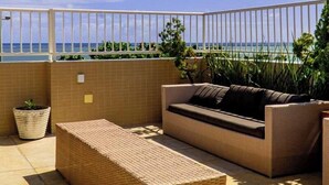 Eine Dachterrasse mit Rattan-Sofa und passendem Hocker, einer Topfpflanze und Blick auf den Ozean.