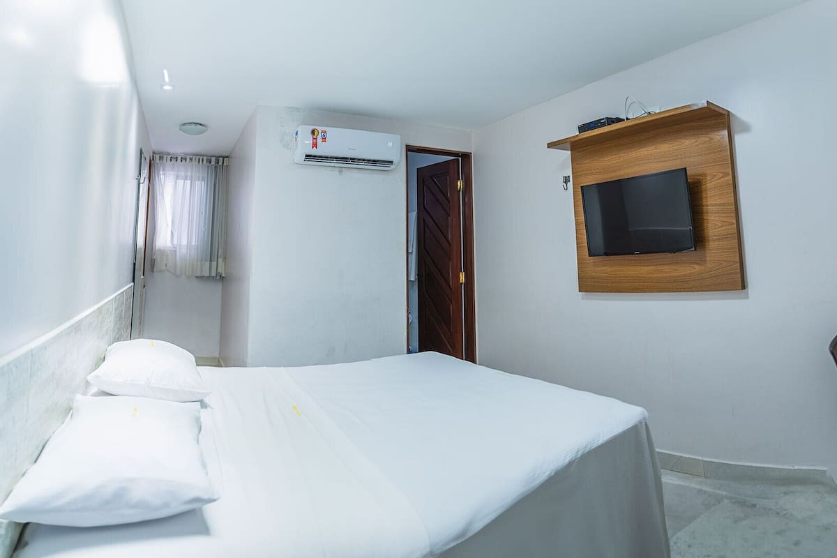 Ein kleiner, sauberer Raum mit einem Bett, einem an der Wand montierten Fernseher und einer Klimaanlage.