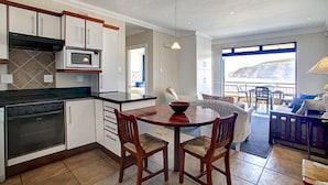 A modern kitchen with white cabinets, a microwave, and a stove. There is a dining area with a wooden table and chairs. - Makarios on Sea (Herolds Bay)