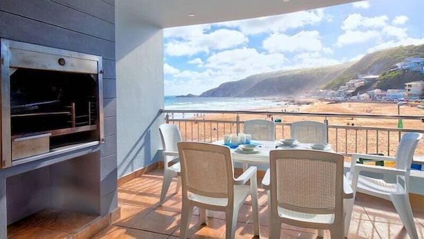 A balcony with a grill, dining table, and chairs overlooking a beach. - Makarios on Sea (Herolds Bay)