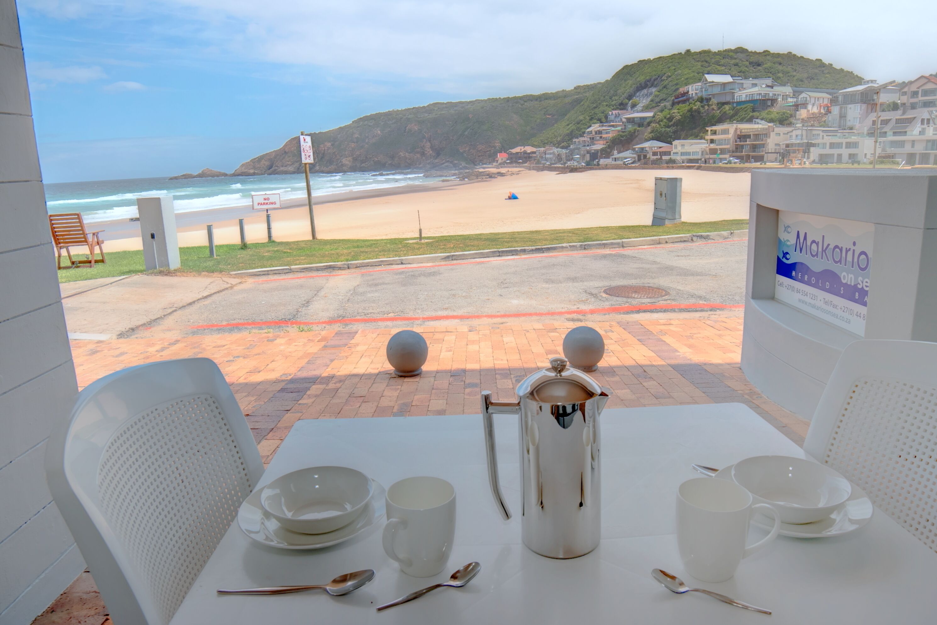 A table set with white plates, cups, and a coffee pot, overlooking a beach with a sign reading "Makarios."