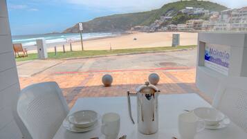 A table set with white plates, cups, and a coffee pot, overlooking a beach with a sign reading "Makarios."