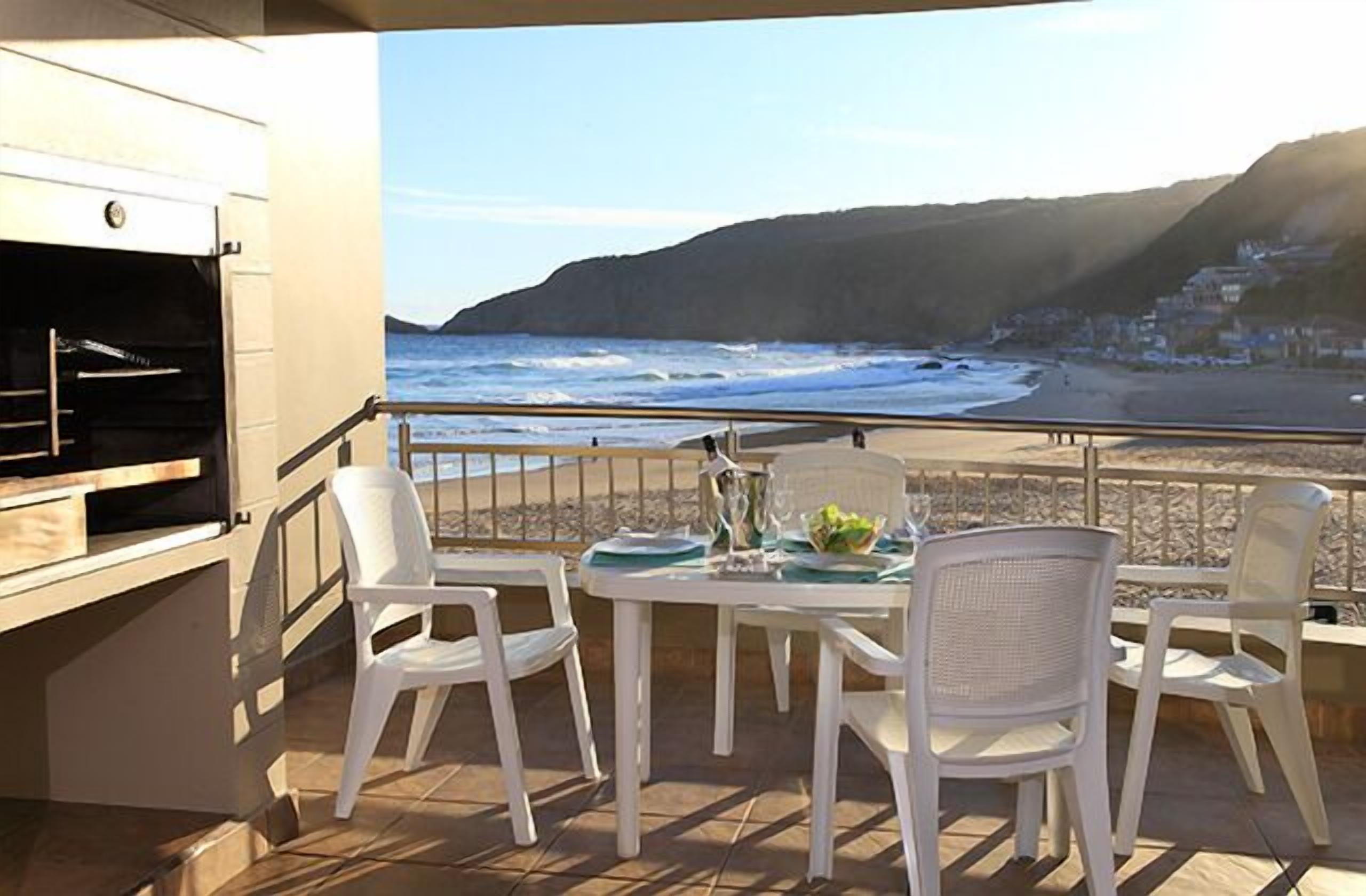 A balcony with a table set for two, overlooking a beach with waves and a mountain in the background.