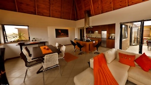 A modern living room with a dining area, a kitchen, and a large window with a view. - Moringa Gardens (Swakopmund)