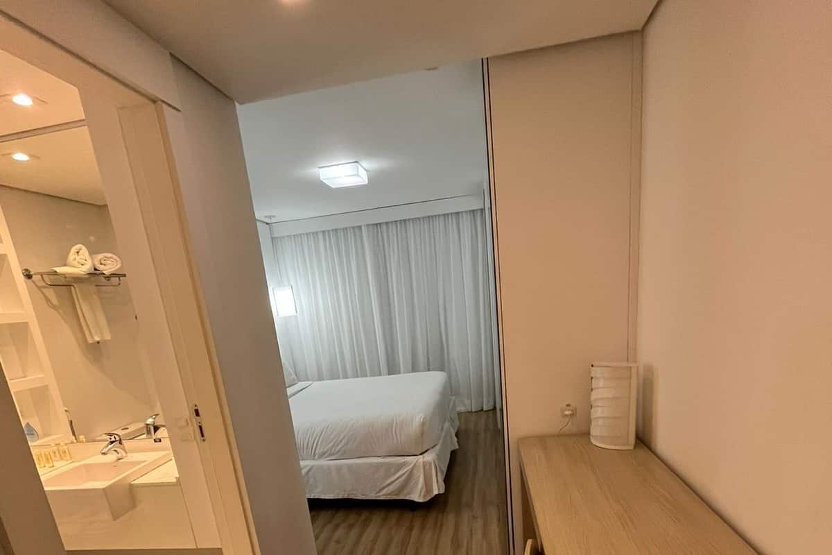 Een hotelkamer met een houten vloer, een bed met witte lakens, een nachtkastje, een spiegel en een badkamer met een wastafel en handdoekhouder.