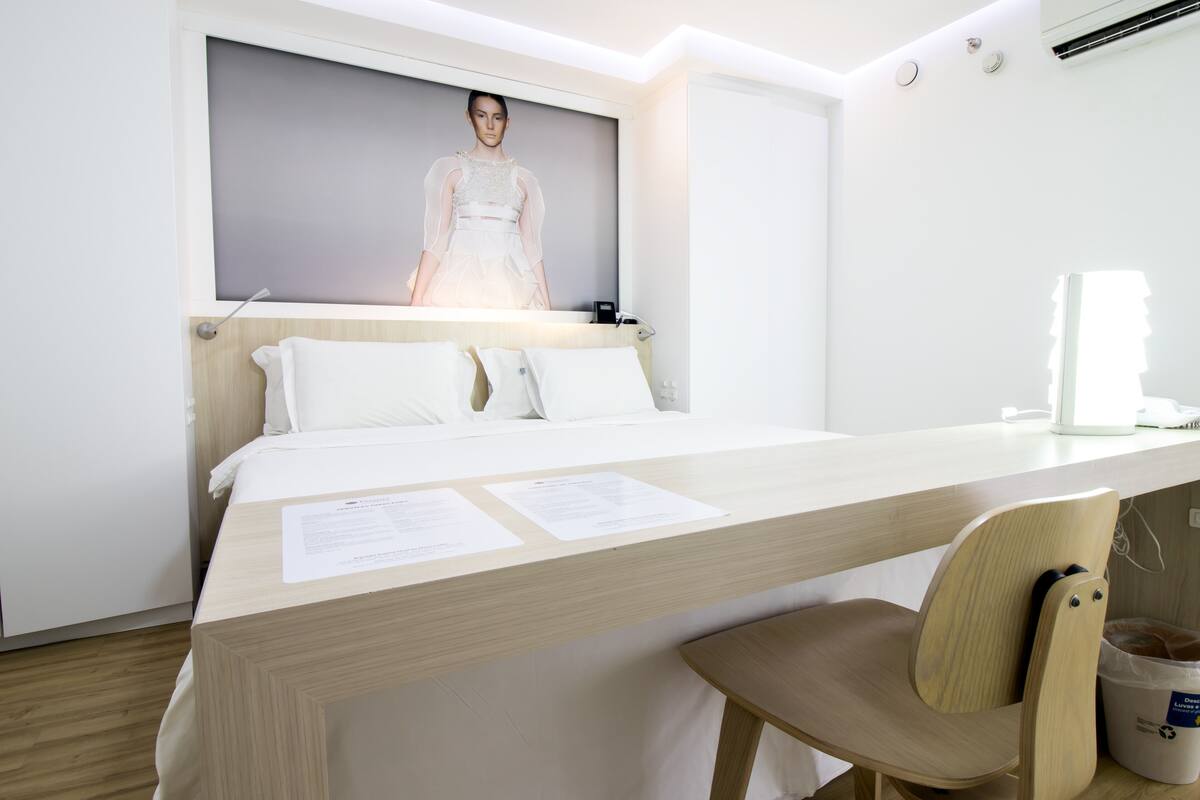 Een moderne hotelkamer met een groot bed, een bureau met papieren, een stoel en een wandtelevisie waarop een model te zien is.