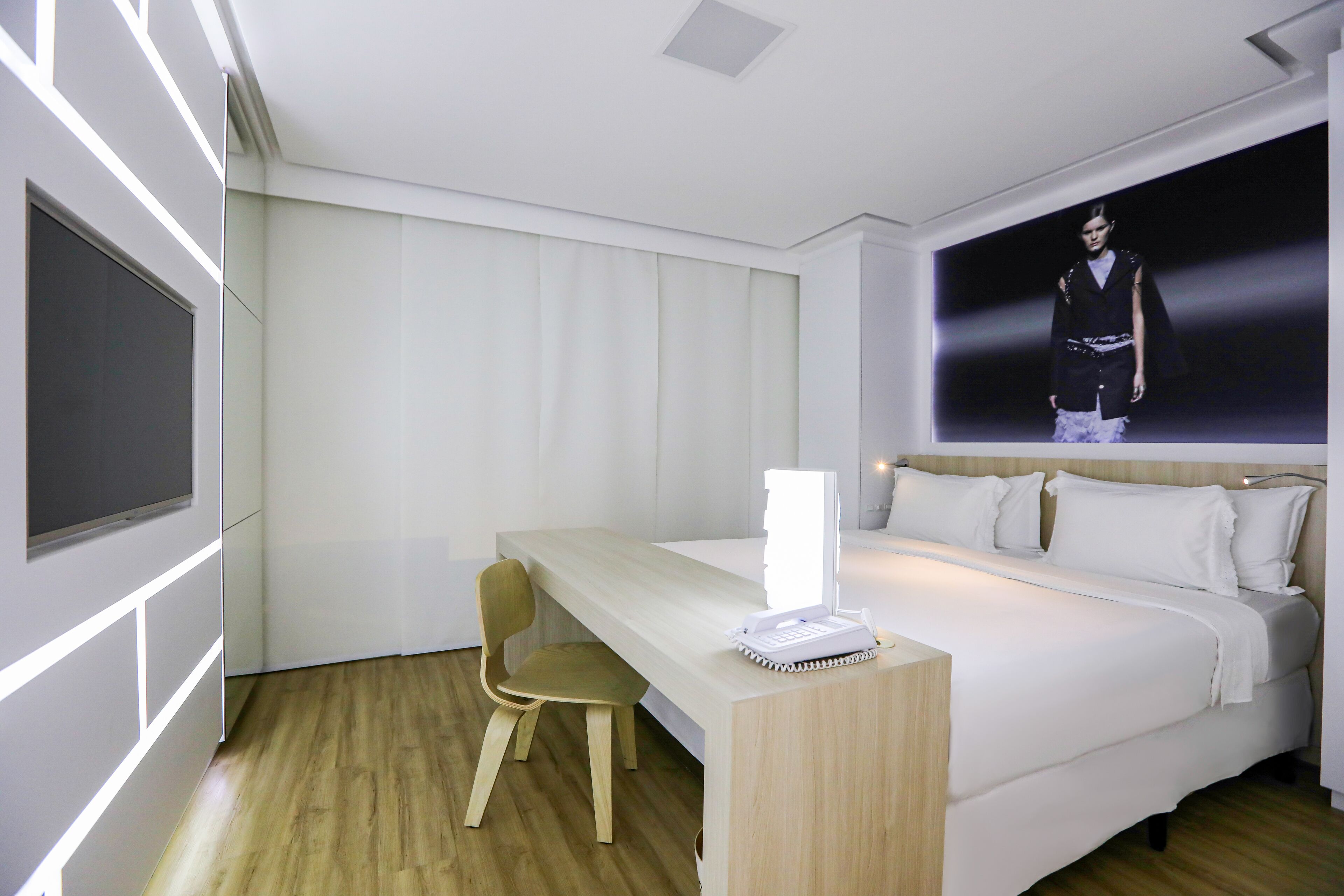 A modern hotel room with a bed, a desk, a chair, and a large screen displaying a person.