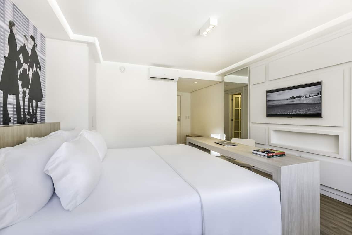 Una habitación de hotel moderna con una cama grande, un escritorio con una computadora portátil, un televisor de pantalla plana y un cuadro en la pared.