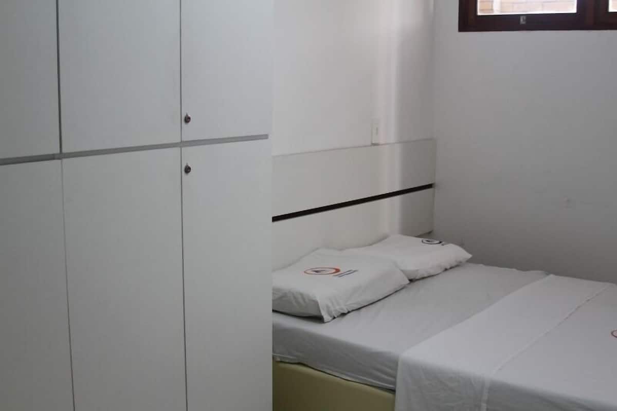 A small, single-bed room with a white bed, a white wardrobe, and a window with blinds.