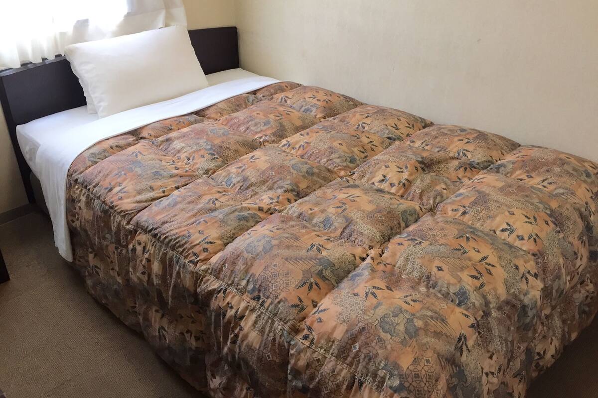 A neatly made bed with a patterned bedspread and white pillows, a dark headboard, and a window with sheer curtains.
