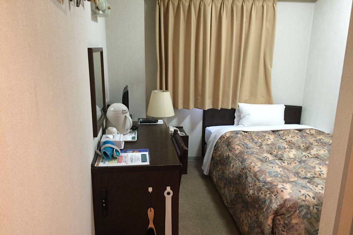 A hotel room with a bed, a desk with a lamp, a trash bin, and a mirror.