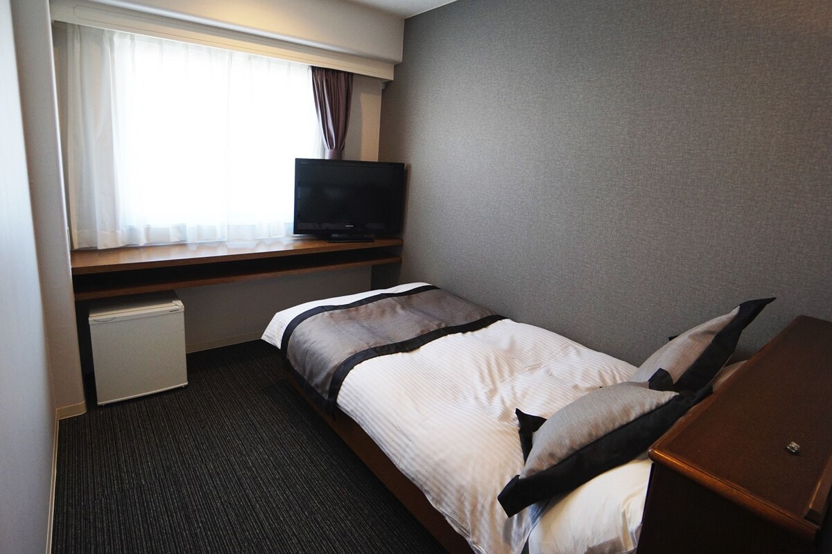 A hotel room with a bed, a television on a wooden shelf, a small desk, and a window with curtains.