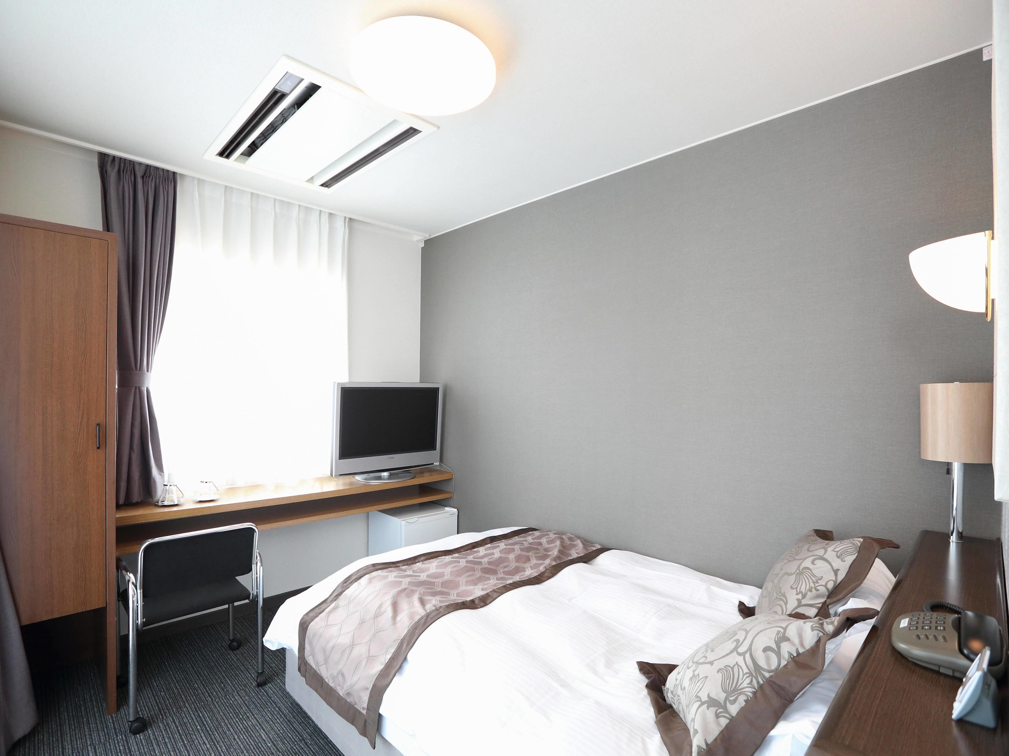 A hotel room with a bed, a desk with a computer, a chair, a TV, a desk lamp, and a telephone.
