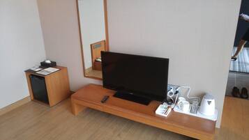 Habitació d'hotel amb un prestatge de fusta per a la televisió, un televisor de pantalla plana, un escriptori petit amb un telèfon, un mirall i una bandeja amb una cafetera, tasses i vasos.