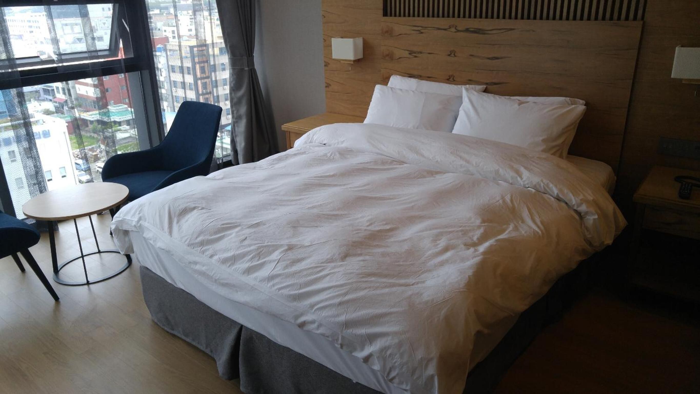 A hotel room with a large bed, a small round table, a chair, and a view of the city through the window.