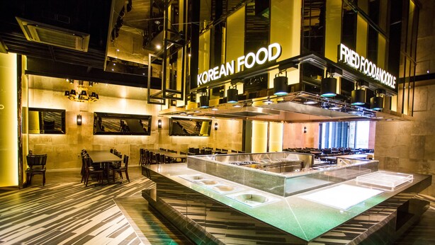 A modern Korean restaurant with a central island serving station, multiple seating areas, and a visible kitchen area. - J Square Hotel (Jinju)