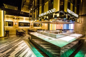 A modern Korean restaurant with a central island serving station, multiple seating areas, and a visible kitchen area. - J Square Hotel (Jinju)