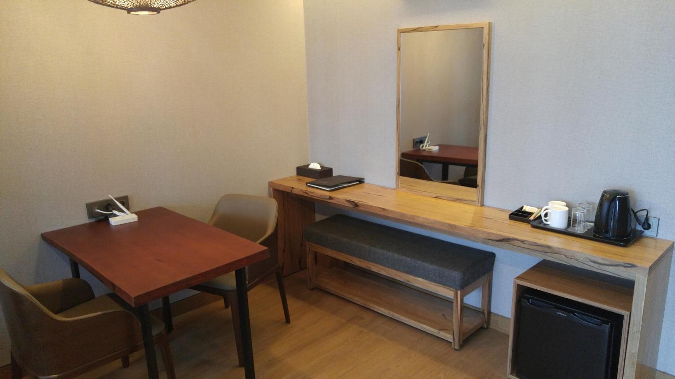 Habitación de hotel compacta con un escritorio de madera, un banco, una pequeña zona de café y una kitchenette integrada.