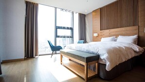 A modern hotel room with a large bed, a bench, a chair, and a view of the city through large windows. - J Square Hotel (Jinju)