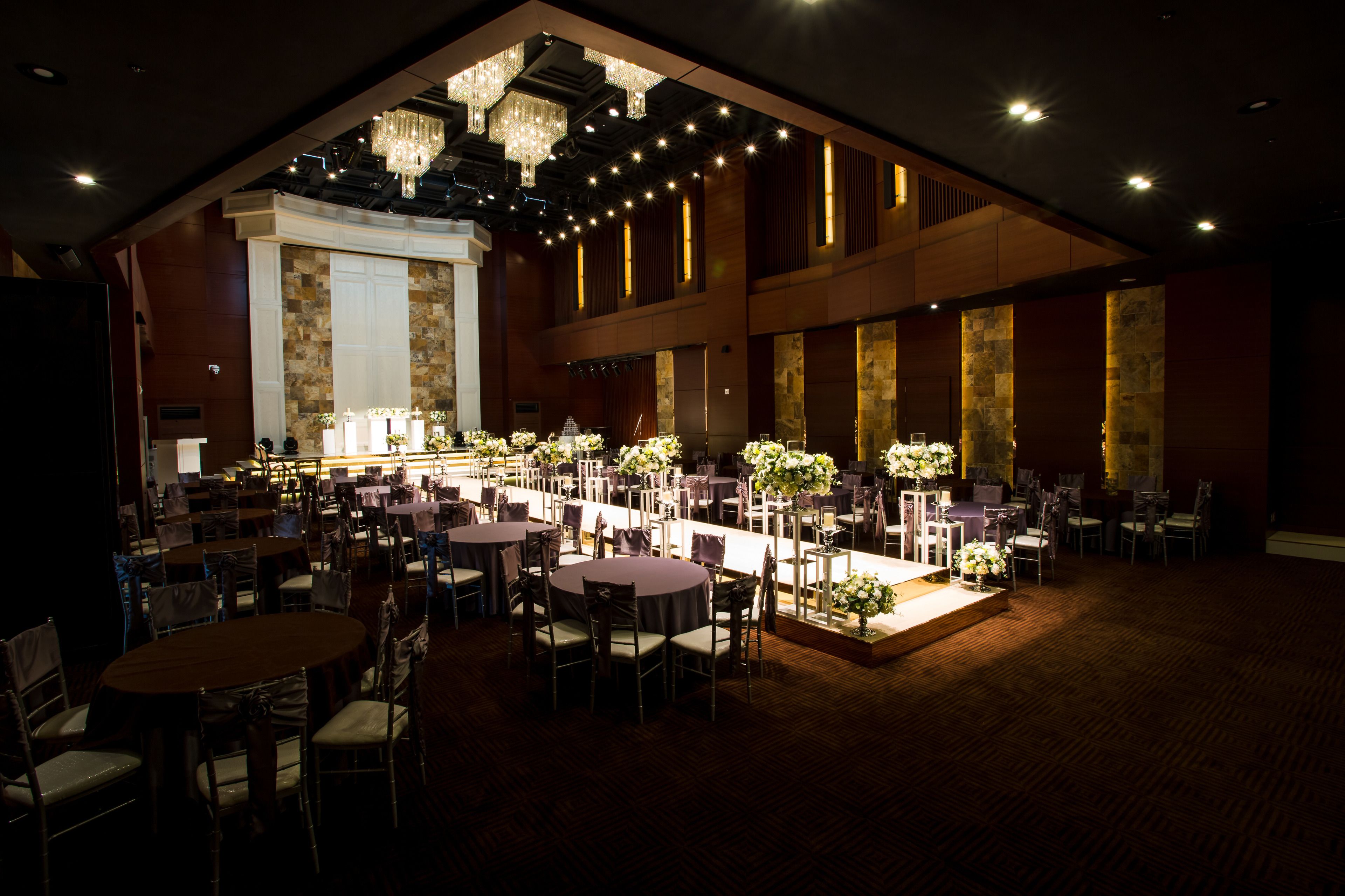 A spacious banquet hall with multiple round tables and chairs, a stage with a backdrop, and a chandelier.