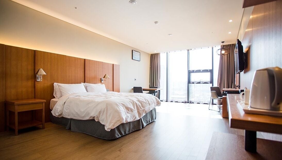 A hotel room with a bed, bedside tables, a desk, a chair, a TV, and a large window.