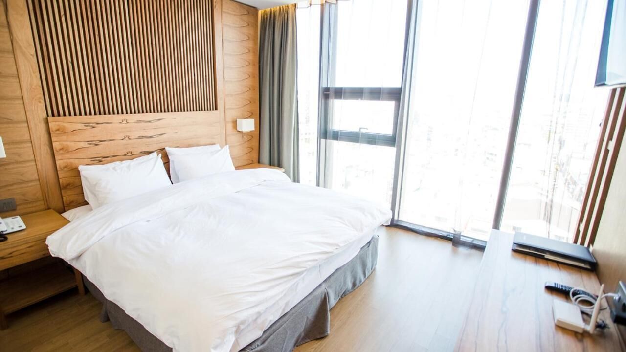 A modern hotel room with a wooden headboard, a large bed with white linens, a bedside table with a lamp, and a view through large windows.