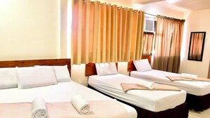 Habitación de hotel con tres camas, cada una con ropa blanca y una toalla rosa. La habitación tiene un cabecero de madera, cortinas color beige y un cuadro en la pared.