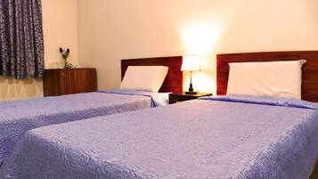 A hotel room with two single beds, a nightstand with a lamp, a wooden headboard, and a window with curtains.