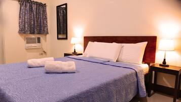 A neatly made bed with a blue bedspread and white pillows, two bedside lamps, a wall-mounted air conditioner, and a window with curtains.