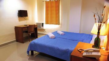 Habitación de hotel con cama, escritorio, silla, televisor, lámpara y teléfono sobre una mesita de noche.