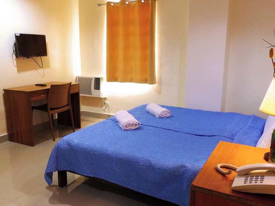 Habitación de hotel con cama, escritorio, silla, televisor, lámpara y teléfono sobre una mesita de noche.