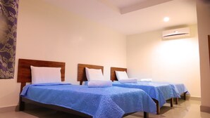 Habitación de hotel con tres camas, cada una con cubrecama azul y almohadas blancas. Cada cama tiene un cabecero de madera, y hay un equipo de aire acondicionado montado en la pared.