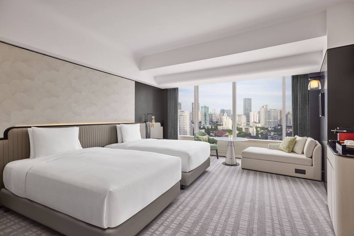 A modern hotel room with two beds, a sofa, and a city view.