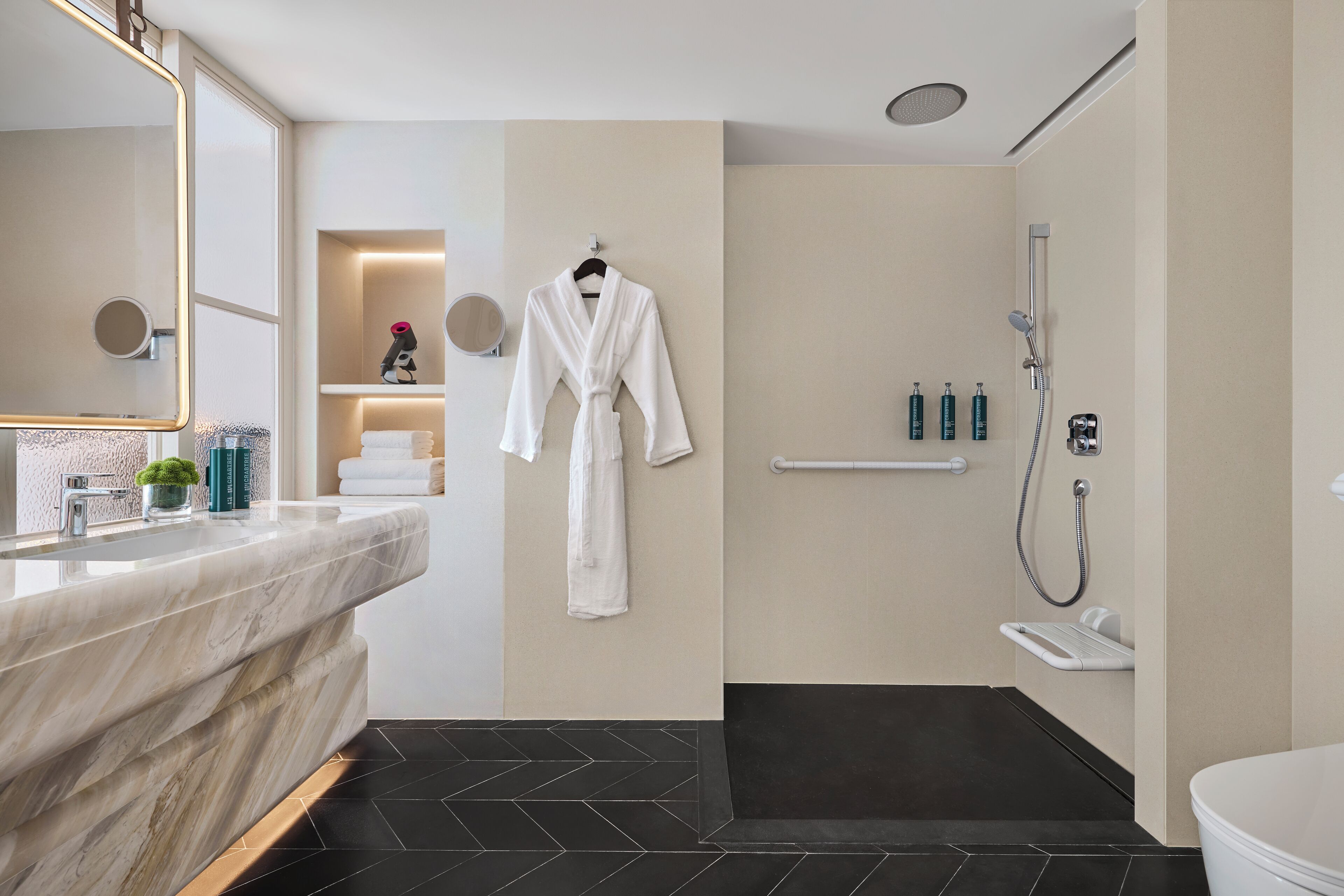 A modern bathroom with a marble vanity, a robe hanging on the wall, a shower area, and a small shelf with toiletries.