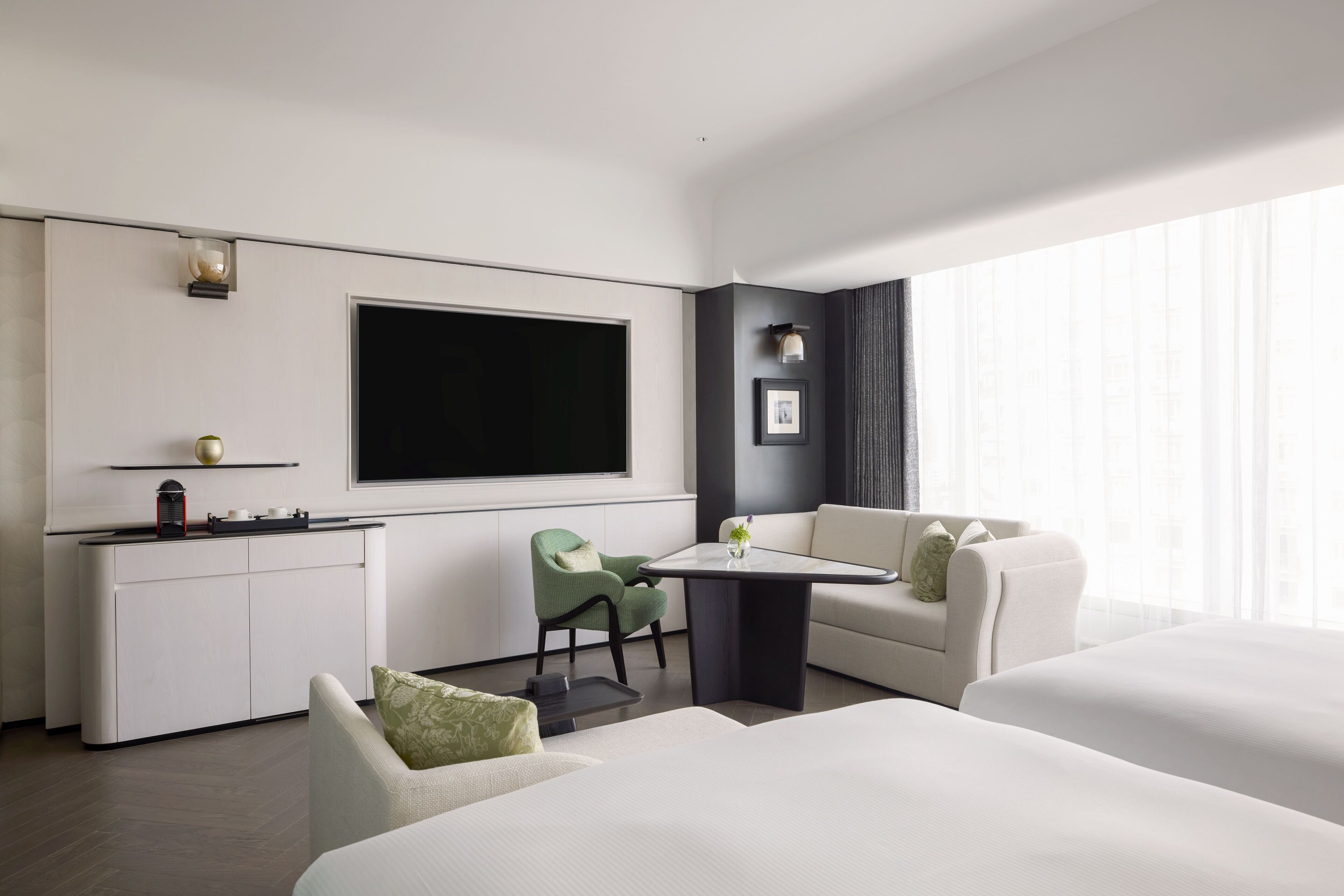 A modern hotel room with a flat-screen TV, a small kitchenette, a sofa, a dining table, and a large window providing natural light.